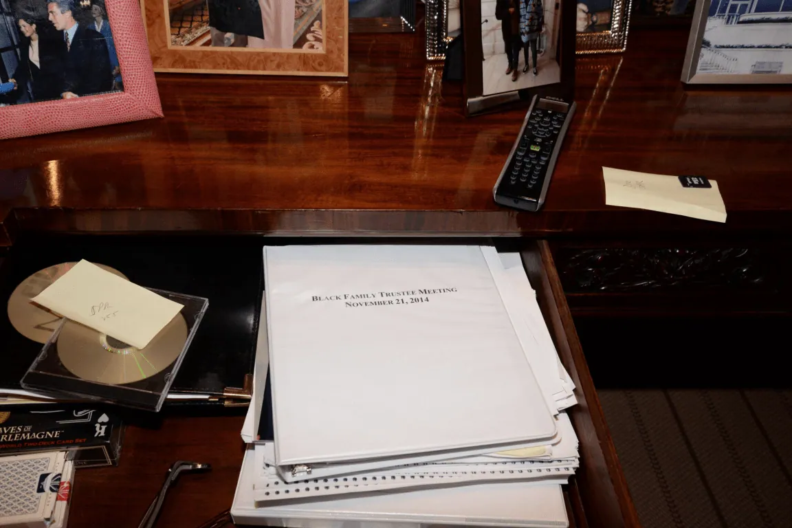 EFTA00001347 - Wooden desk holds a stack of documents from the Black Family Trustee Meeting alongside photos a remote control and a CD case