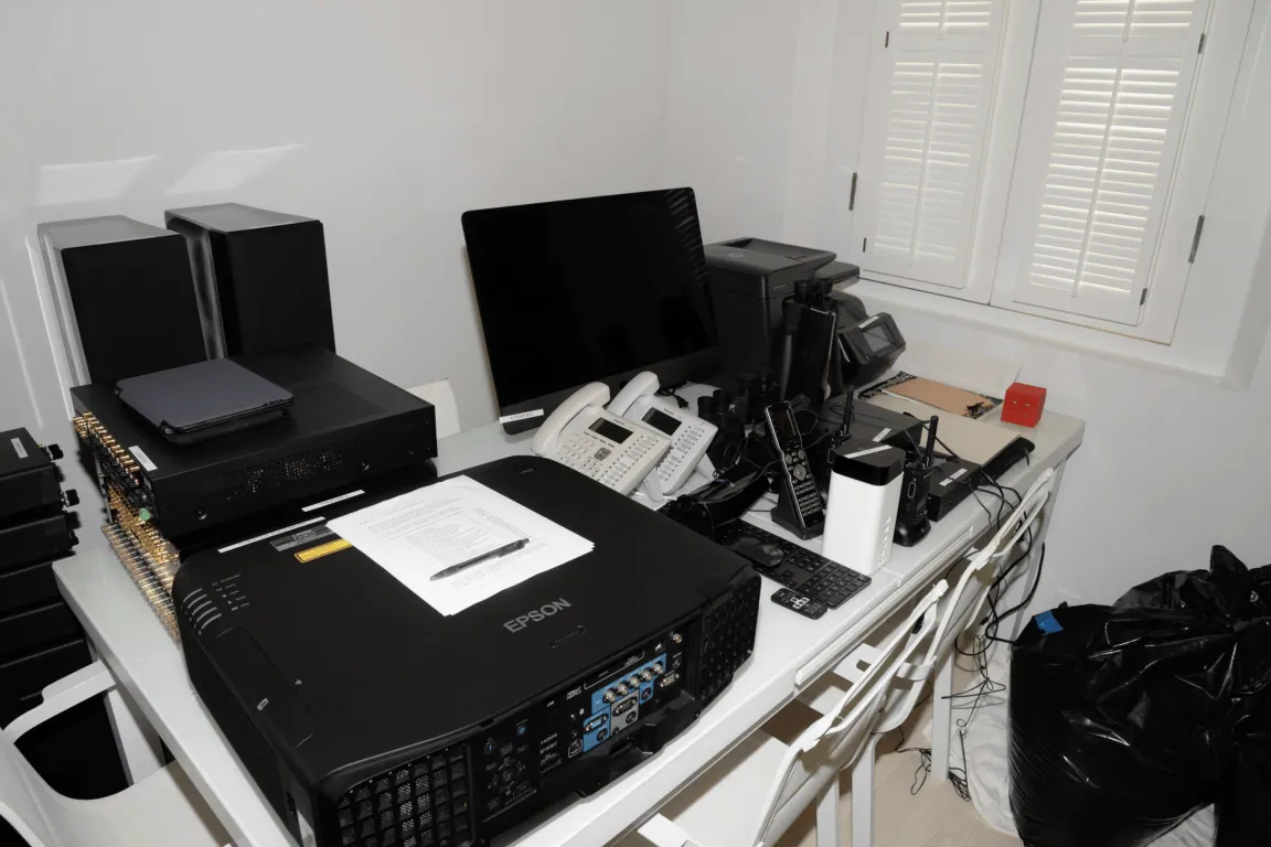 EFTA00001583 - Cluttered office desk with an Epson projector multiple electronic devices a computer monitor and various cables next to a window with shutters