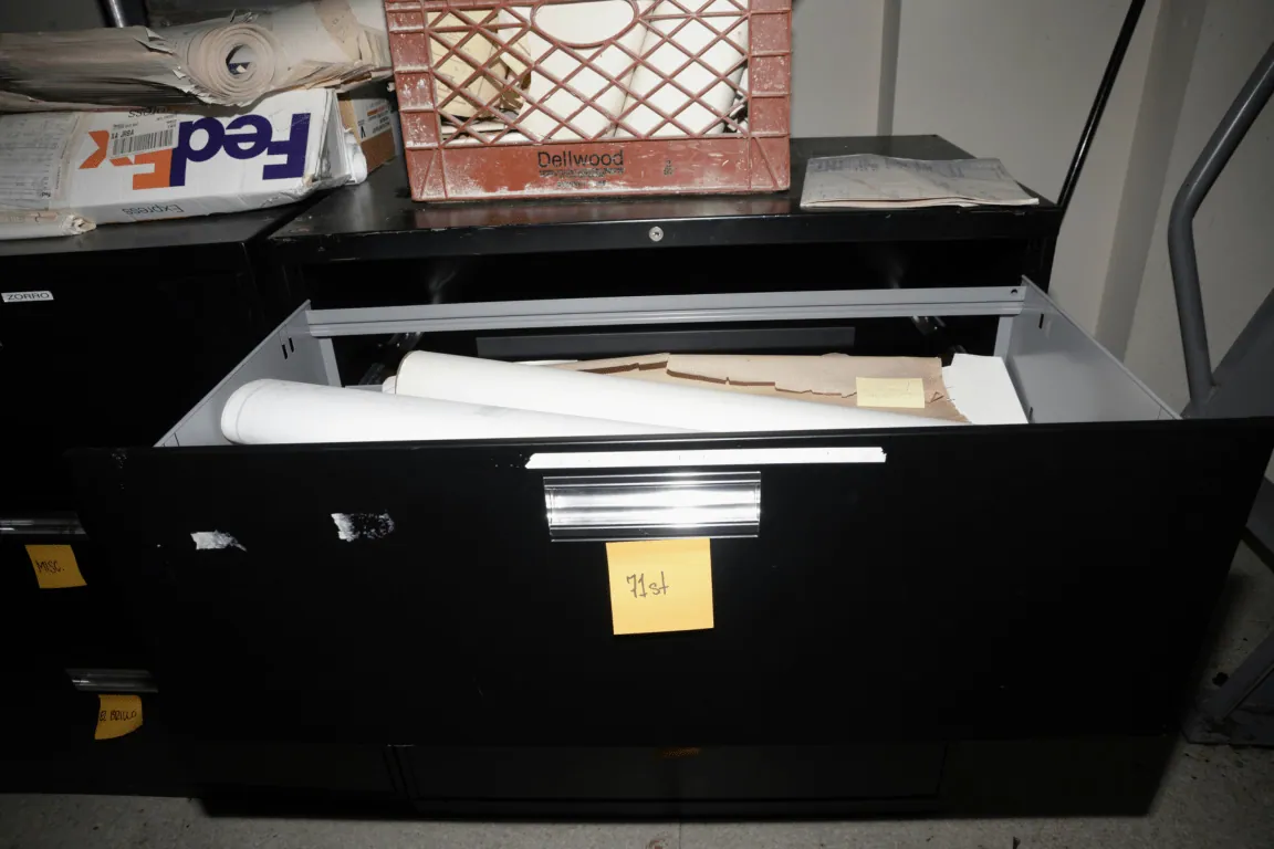 EFTA00000040 - Black filing cabinet drawer open revealing rolled white paper and a yellow sticky note labeled List with a red milk crate and FedEx box on top