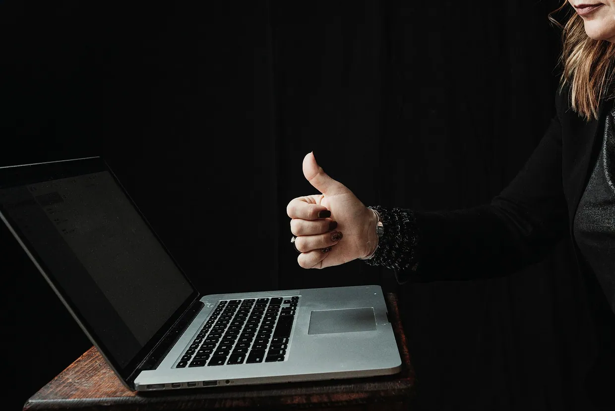 Person giving thumbs up gesture while using laptop against dark background in a home office setting